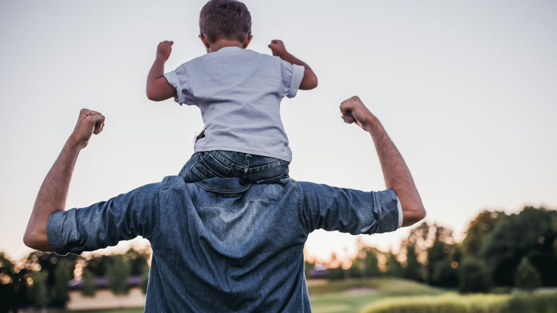 A father carrying his young son on his shoulders, both flexing their arms triumphantly outdoors